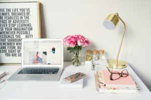 A bright, aesthetic workspace flat-lay featuring a laptop, a gold desk lamp, a bouquet of pink roses in a mason jar, and an inspirational framed quote on a white desk.